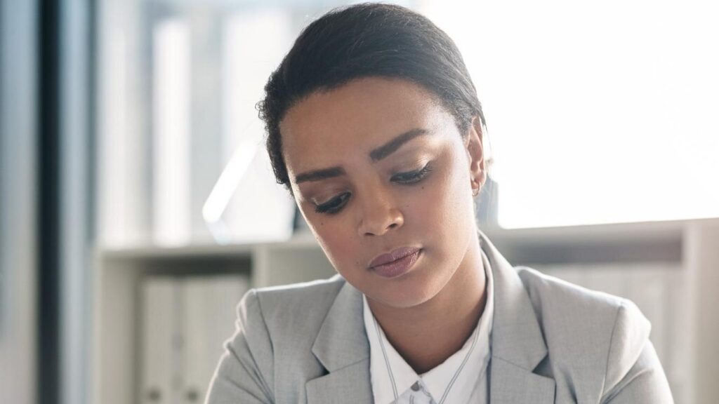 A professional in a grey blazer looking down in thought at a bright office desk